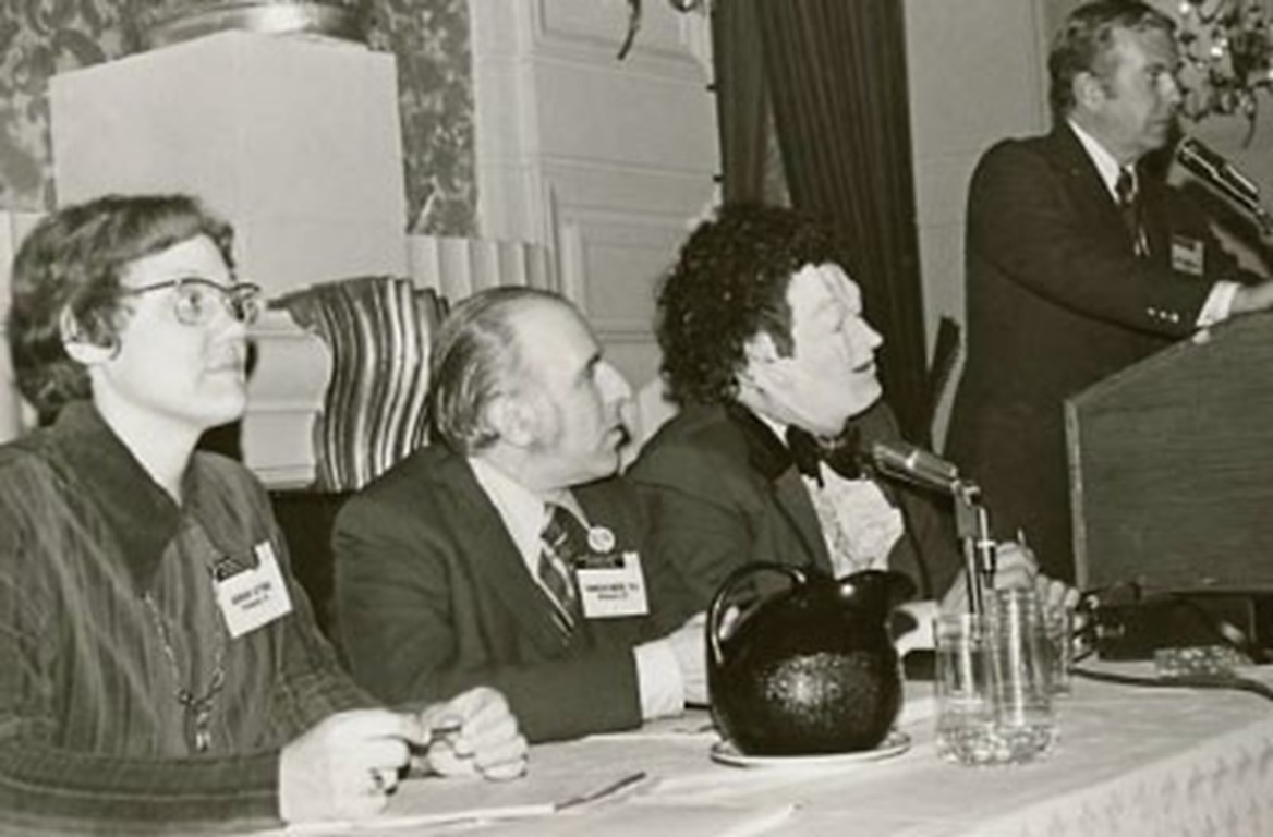 people sitting behind a table with microphones. One person standing at a podium speaking